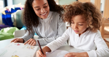 A woman pointing at a book while observing a little girl writing