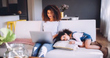 Young female child lying next to mother while she works on laptop