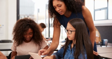 Teacher of color assisting two girl students of color in a classroom