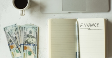 Money, notebook with 'finance' written on page, coffee, and laptop on a desk