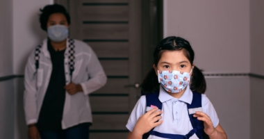 Child wearing a face mask and backpack while mother stands behind her with a face mask on